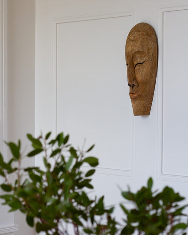 Vintage wooden face sculpture on a white wall with greenery in the foreground, curated by Parliament Interiors in Vancouver, BC.