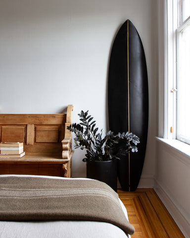 Black wooden surfboard leaning against a white wall next to intricately carved vintage wooden bench, in front of a bed with a vintage wool blanket and white linens and potted plant in a heritage house with original wood flooring.