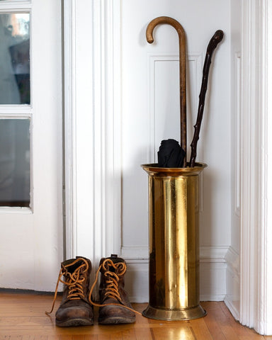 Vintage brass umbrella stand with a cane and black umbrella next to a pair of brown boots on a wooden floor. Curated by Parliament Interiors in Vancouver, BC.