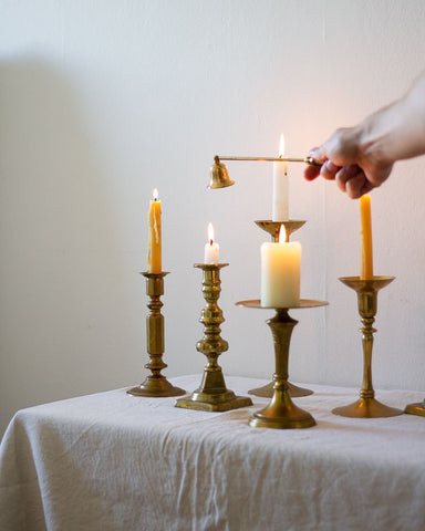 Five vintage brass candle holders, each with a lit candlestick on a white tablecloth in front of a white wall. A hand reaches into the frame with a vintage brass candle snuffer.